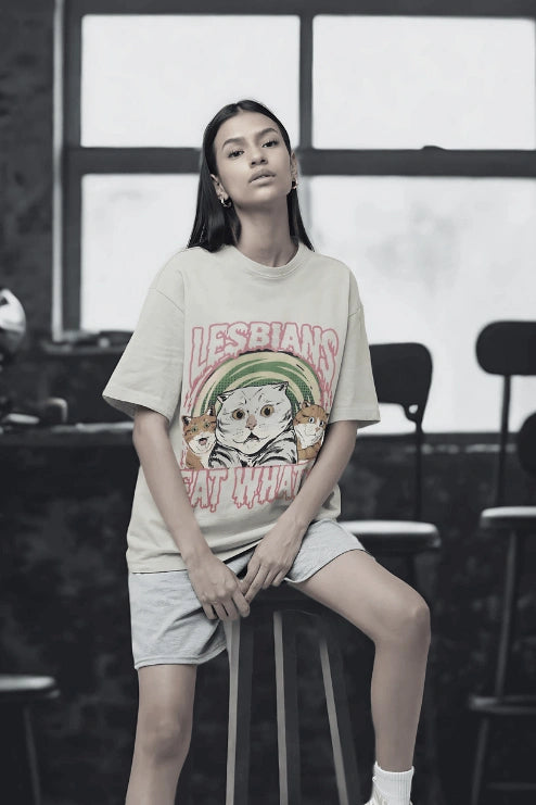 Person wearing a lesbians eat what unisex graphic oversized  t-shirt sitting on a stool indoors.